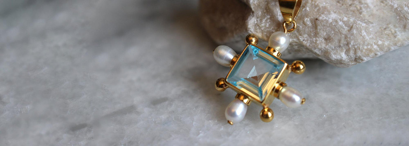 a Blue Topaz Cross Pendant from Freya Rose's Something Blue Collection laid upon a white stone.